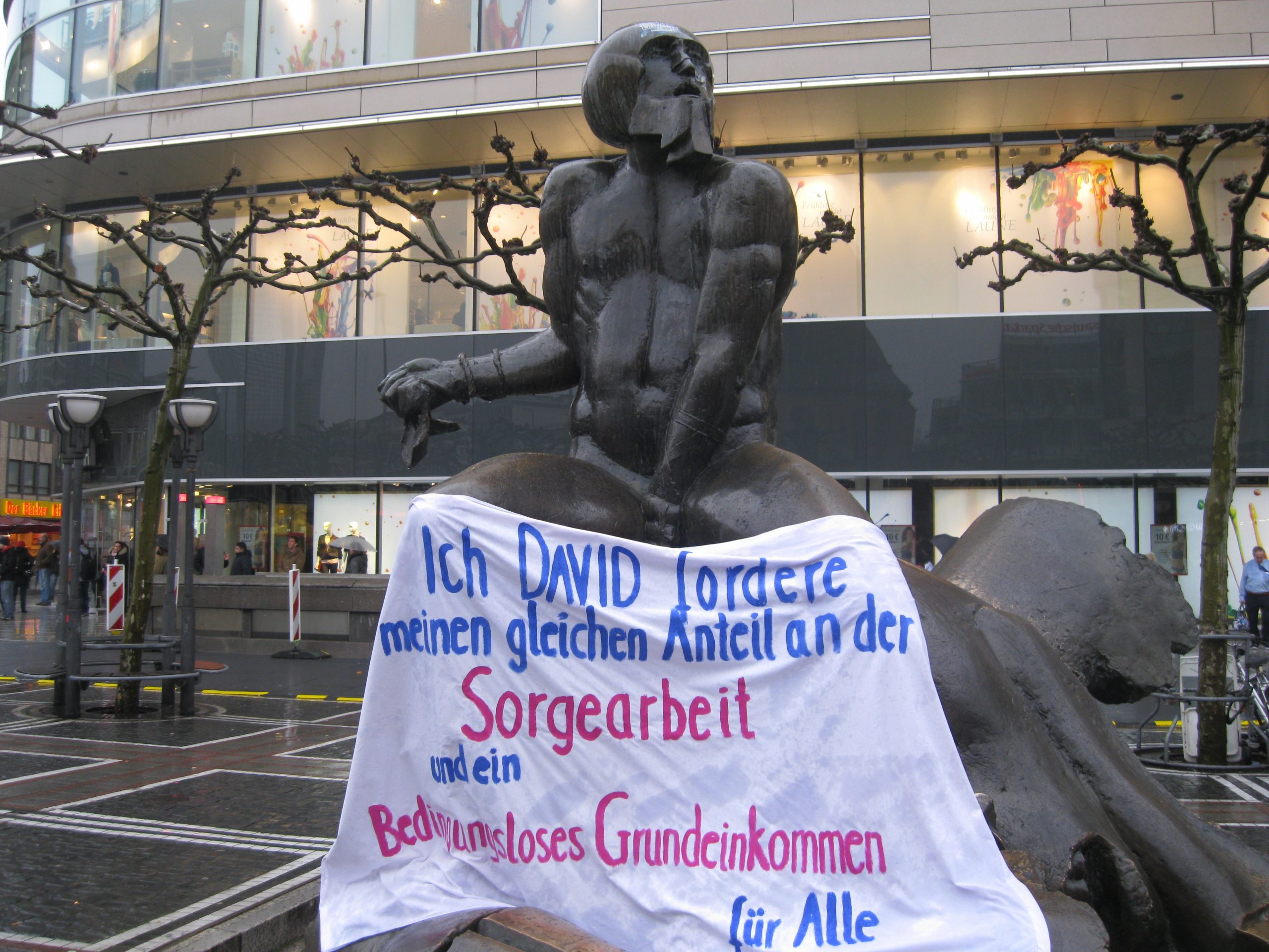 David (und Goliath-)Statue auf der Zeil mit Schuerze auf der steht: Ich foderer die Haelfte der Sorgearbeit und ein BGE fuer alle